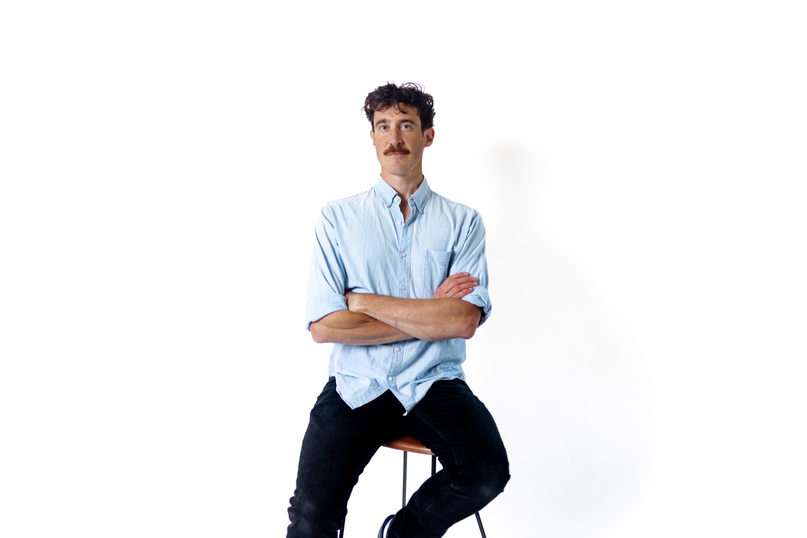 Portrait of Alec Jankowski, seated on a stool with his arms crossed, looking at the camera.